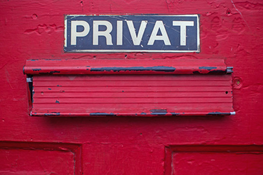 Großaufnahme Briefschlitz in rot lackierter Haustüre, darüber "Privat"-Schild, alles sieht sehr benutzt aus